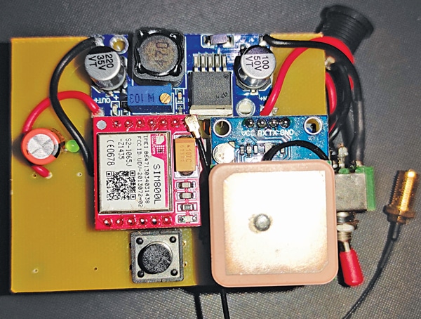 Emergency SOS hardware prototype on a perfboard: blue buck regulator board with capacitors and inductor, red GSM module with metal shield, ceramic GPS patch antenna, tactile push button and toggle switch